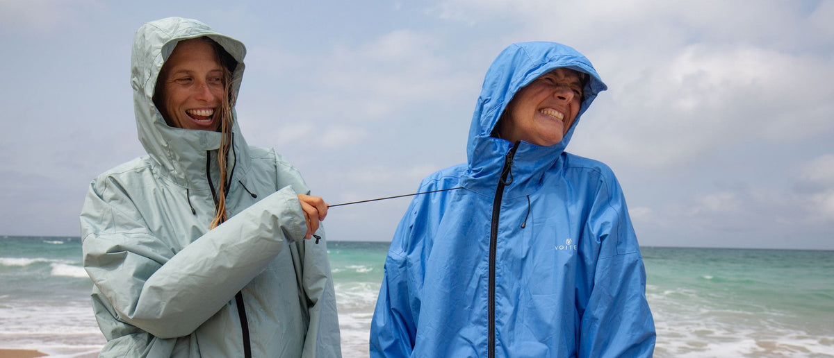 Deux personnes portant des vestes de pluie légères à capuche - Allweather Coat - se tiennent sur une plage, riant pendant que l'une tire sur le cordon de serrage de la veste de l'autre. La veste de gauche est vert menthe pâle et celle de droite est bleu moyen.
