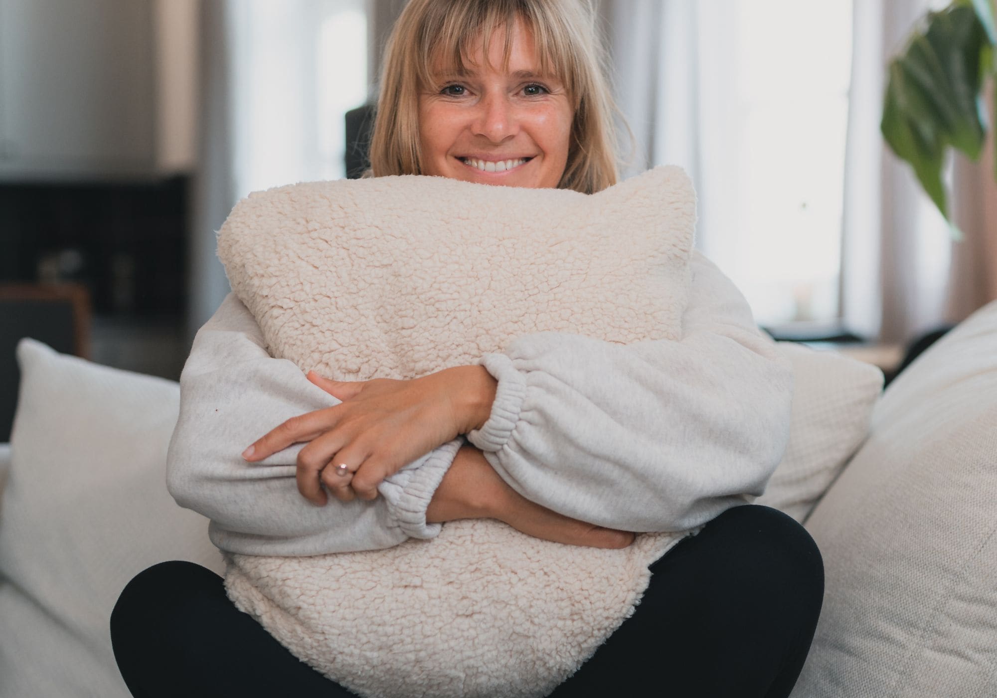 Femme souriante assise sur un canapé, serrant dans ses bras un grand coussin moelleux en peluche crème VOITED, ambiance intérieure chaleureuse et lumineuse-Cloudtouch