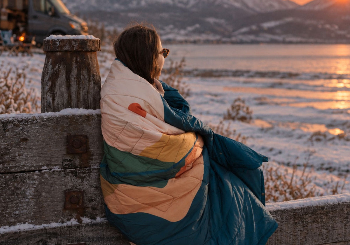 Femme enveloppée dans une couverture VOITED CloudTouch, adossée à une barrière enneigée face à un lac au coucher du soleil. AI modified