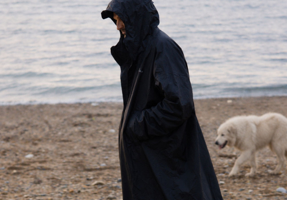 Homme en VOITED Allweather Coat noir se promenant sur la plage, accompagné d’un chien blanc. AI modified
