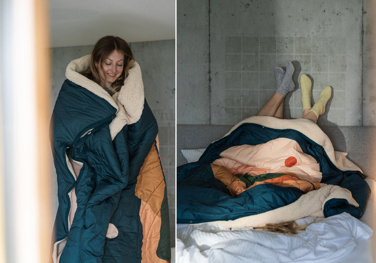 Femme enveloppée dans une couverture VOITED Cloudtouch doublée de sherpa ; deux personnes allongées sous la couverture sur un lit, pieds en chaussettes contre un mur en béton.