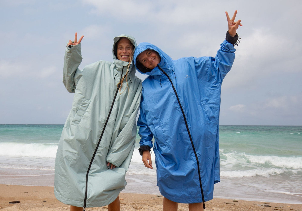 Deux femmes souriantes portant des VOITED Allweather Coats vert sauge et bleu, capuche sur la tête, faisant le signe de la paix sur une plage face aux vagues.
