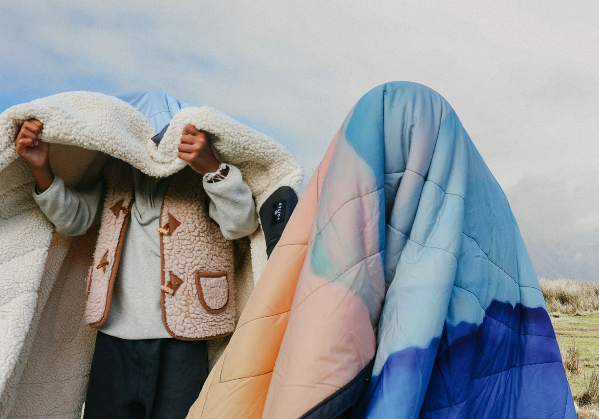 Deux personnes en plein air sous un ciel nuageux. L’une s’enveloppe dans une couverture moelleuse en sherpa, l’autre disparaît entièrement sous une couverture matelassée Voited Cloudtouch au dégradé pêche, bleu clair et bleu profond