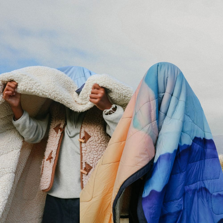 Deux personnes en plein air sous un ciel nuageux. L’une s’enveloppe dans une couverture moelleuse en sherpa, l’autre disparaît entièrement sous une couverture matelassée Voited Cloudtouch au dégradé pêche, bleu clair et bleu profond