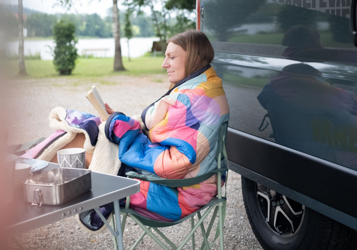 Femme enveloppée dans la couverture VOITED Cloudtouch (blocs de couleurs) lisant tranquillement en camping au bord d'un lac