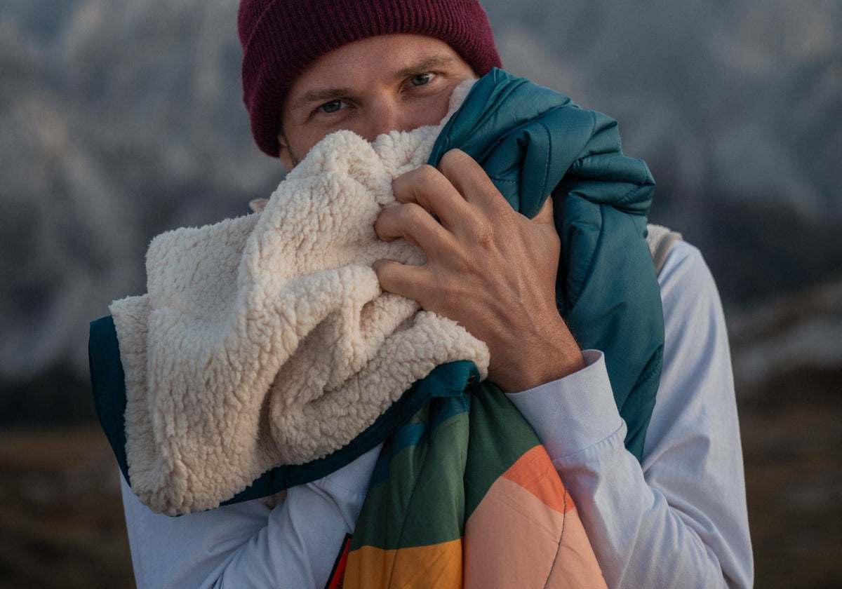 Homme avec un bonnet tenant une couverture VOITED Cloudtouch avec un côté sherpa crème et un côté turquoise et orange, montagnes en arrière-plan.