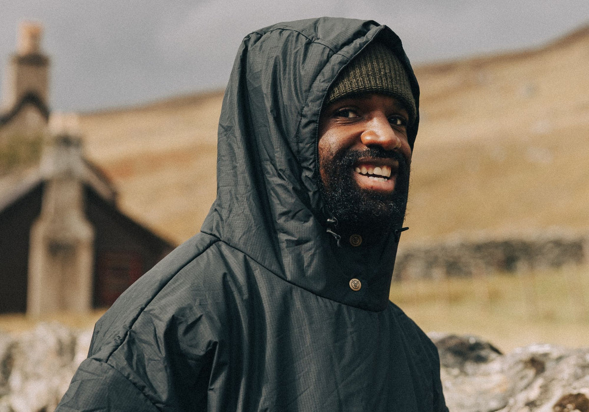 Homme souriant portant un poncho VOITED vert foncé, capuche relevée sur un bonnet, devant un paysage vallonné et une maison en pierre.