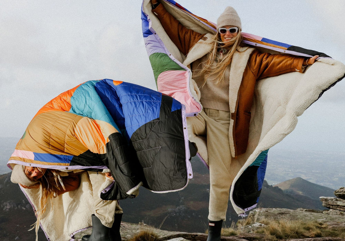 Offre groupée Value Set VOITED Cloudtouch : deux femmes au sommet d’une montagne avec deux couvertures Cloudtouch au motif patchwork coloré et doublure sherpa, flottant au vent.