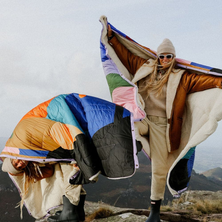 Offre groupée Value Set VOITED Cloudtouch : deux femmes au sommet d’une montagne avec deux couvertures Cloudtouch au motif patchwork coloré et doublure sherpa, flottant au vent.