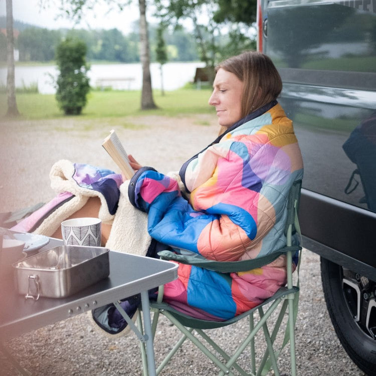 Femme enveloppée dans la couverture VOITED Cloudtouch (blocs de couleurs) lisant tranquillement en camping au bord d'un lac