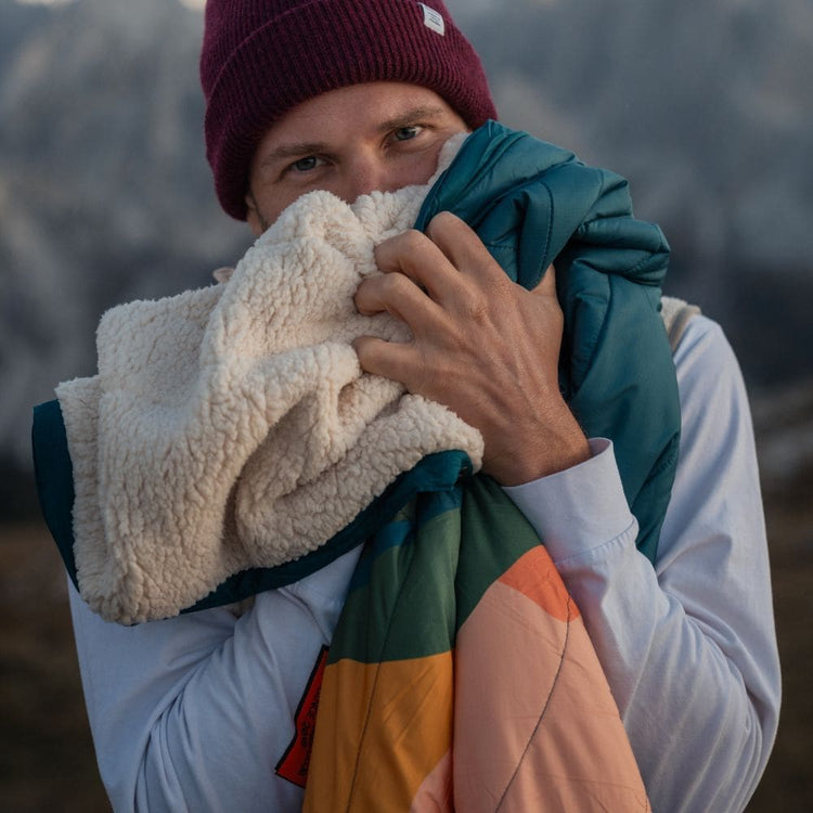 Homme avec un bonnet tenant une couverture VOITED Cloudtouch avec un côté sherpa crème et un côté turquoise et orange, montagnes en arrière-plan.