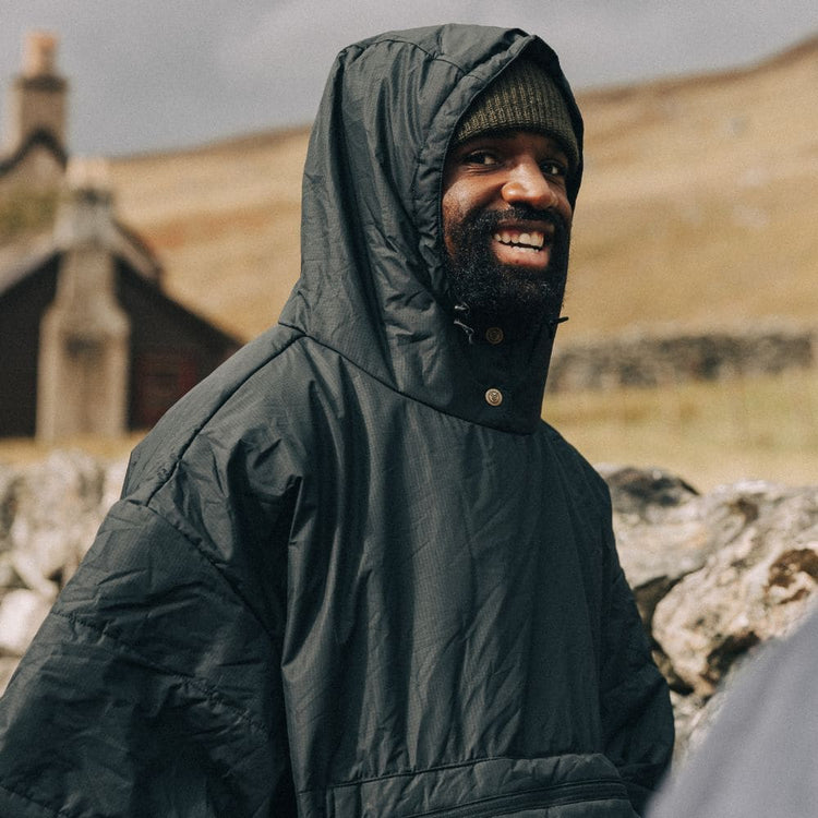 Homme souriant portant un poncho VOITED vert foncé, capuche relevée sur un bonnet, devant un paysage vallonné et une maison en pierre.