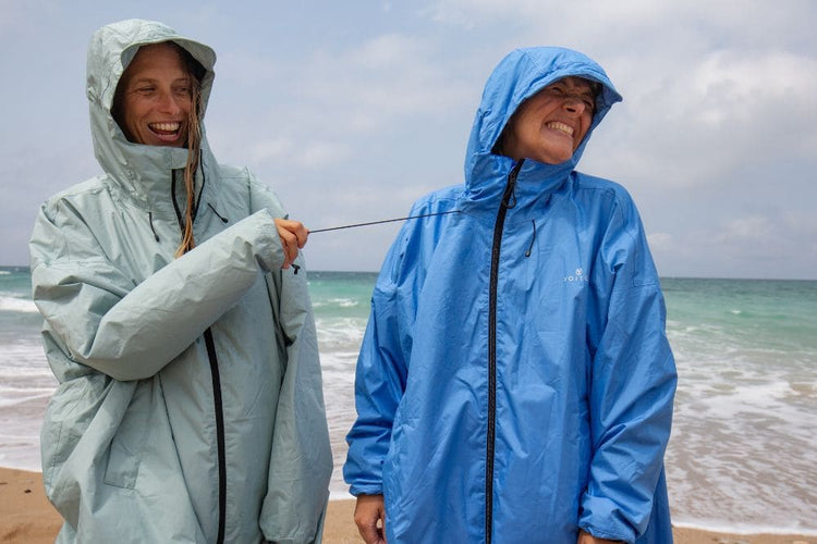Deux personnes portant des vestes de pluie légères à capuche - Allweather Coat - se tiennent sur une plage, riant pendant que l'une tire sur le cordon de serrage de la veste de l'autre. La veste de gauche est vert menthe pâle et celle de droite est bleu moyen.

