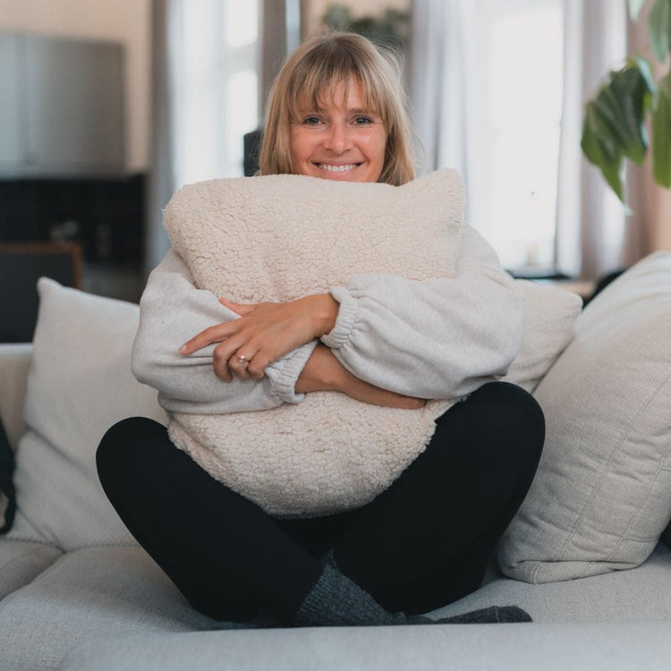 Femme souriante assise sur un canapé, serrant dans ses bras un grand coussin moelleux en peluche crème VOITED, ambiance intérieure chaleureuse et lumineuse-Cloudtouch