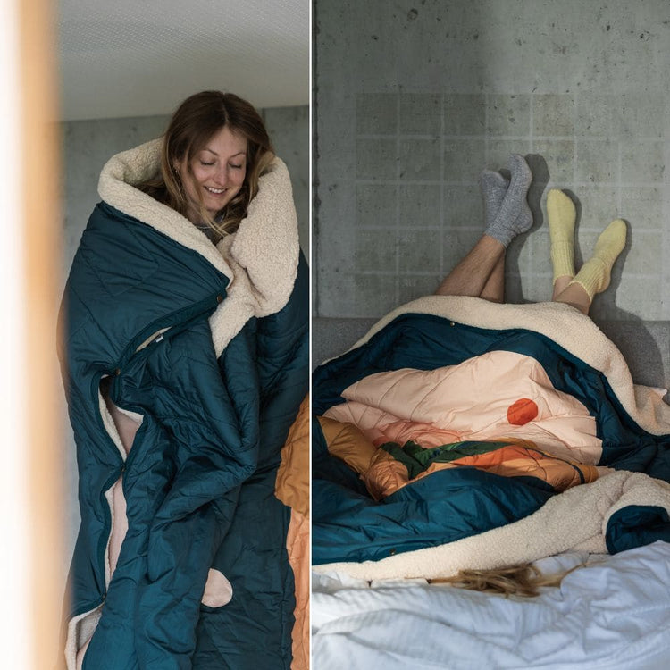 Femme enveloppée dans une couverture VOITED Cloudtouch doublée de sherpa ; deux personnes allongées sous la couverture sur un lit, pieds en chaussettes contre un mur en béton.
