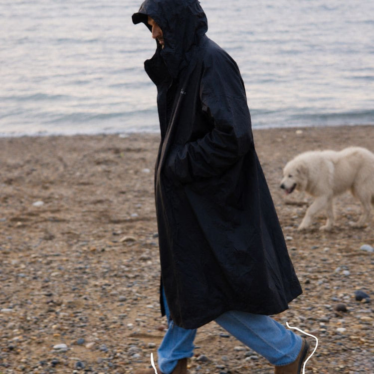 Homme en VOITED Allweather Coat noir se promenant sur la plage, accompagné d’un chien blanc. AI modified