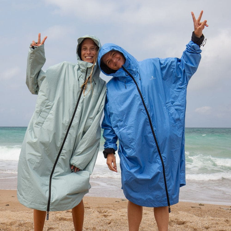 Deux femmes souriantes portant des VOITED Allweather Coats vert sauge et bleu, capuche sur la tête, faisant le signe de la paix sur une plage face aux vagues.